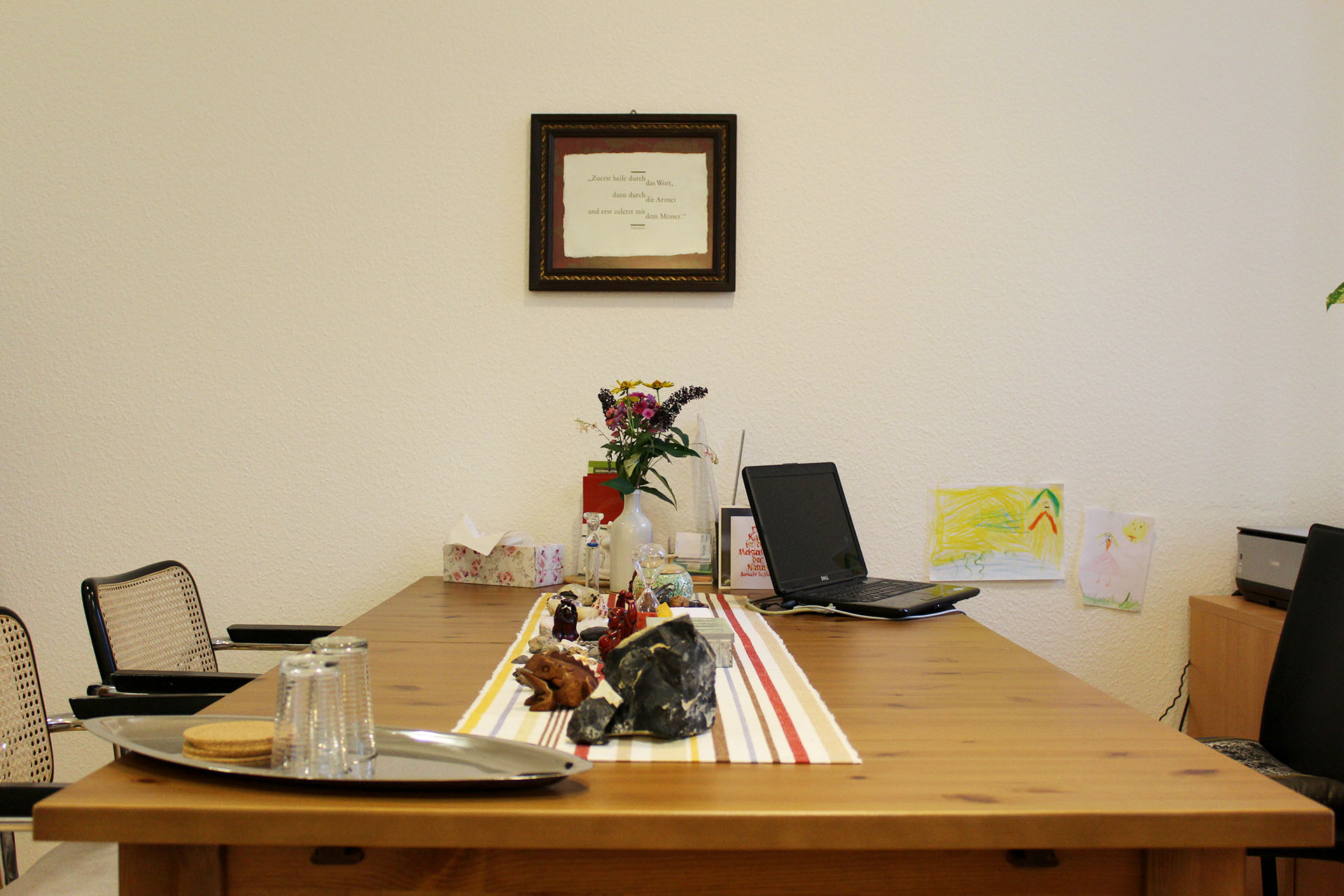 Ein Tisch im Behandlungsraum der Praxis darauf steht der Laptop der Heilpraktikerin, an der Wand hängt ein Bild von einem Zitat von Asklepios.